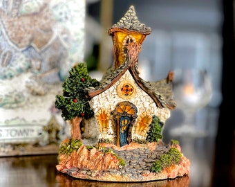 May include: A miniature stone church with a brown roof, a green tree, and a stone pathway leading to the front door. The church is decorated with a stone facade and a small window. The church is sitting on a base of green moss and brown rocks.