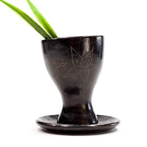 May include: A black ceramic vase with a floral design and a matching saucer. The vase has a wide base and a narrow neck. A green plant is placed inside the vase.