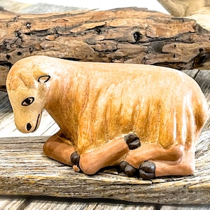May include: A ceramic figurine of a lamb in a resting position. The lamb is primarily tan with darker brown accents on its hooves and face. The figurine is set against a backdrop of weathered wood.