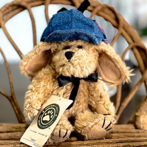 May include: A small, tan teddy bear wearing a blue denim cap and a black bow tie. The bear is sitting on a wooden chair. The bear has a tag that says "INDY" on it.