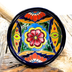 Op de afbeelding: Een kleurrijke keramische kom met een blauwe rand en een bloemenmotief. De kom heeft een centrale bloem met rode bloemblaadjes en een gele kern, omringd door kleinere bloemen en bladeren in tinten groen, geel, oranje en rood.