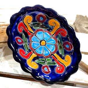 Pode incluir: Um prato de cerâmica azul com borda ondulada apresenta um design floral colorido. O design inclui uma grande flor azul com um centro amarelo, cercada por redemoinhos vermelhos, amarelos e verdes.