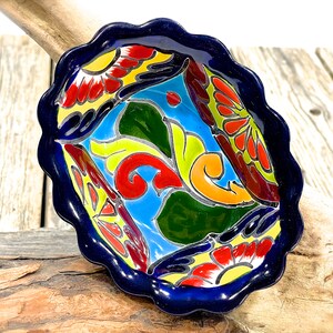 Pode incluir: Uma tigela pequena de cerâmica azul com uma borda ondulada. A tigela é decorada com um padrão floral colorido em tons de vermelho, amarelo, verde e azul.