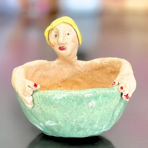 May include: A ceramic figurine of a woman with blonde hair, red fingernails, and a green glazed bowl. The woman is sitting in the bowl with her arms resting on the rim.
