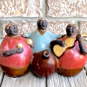 Puede incluir: Tres figuritas de cerámica de personas tocando música. La persona de la izquierda sostiene un objeto pequeño, la persona del medio lleva una camisa azul y la persona de la derecha toca la guitarra.