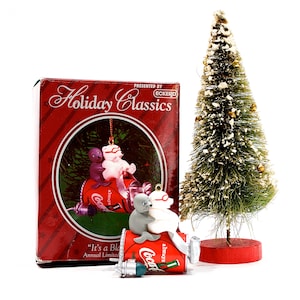 Puede incluir: Una exhibición navideña con una caja roja con el texto "Holiday Classics", un pequeño árbol de Navidad decorado y adornos de Coca-Cola. Los adornos representan focas y osos en una lata de Coca-Cola. La caja dice "It's a Blast".