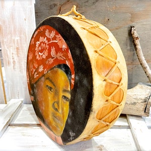 May include: A hand-painted Native American drum with a black background and a portrait of a woman wearing a red headscarf. The drum is made of wood and leather and has a decorative lacing pattern.
