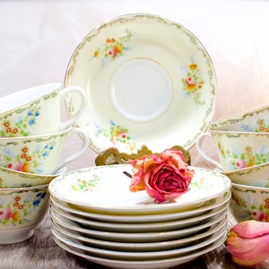 May include: A set of vintage teacups and saucers with a floral pattern in shades of pink, blue, and green. The cups and saucers are stacked on a wooden surface with dried roses.