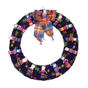 May include: A handmade wreath featuring a dark blue fabric base adorned with small, colorful dolls. The wreath is decorated with a plaid bow in shades of orange, blue, and gray. The dolls are dressed in vibrant, patterned clothing.