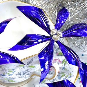 Pu&ograve; includere: Primo piano di un fiocco metallico blu e argento con una tazza da t&egrave; e un piattino bianchi sullo sfondo. Il fiocco &egrave; fatto di strisce sottili e lucide di materiale e ha un aspetto delicato ed elegante.