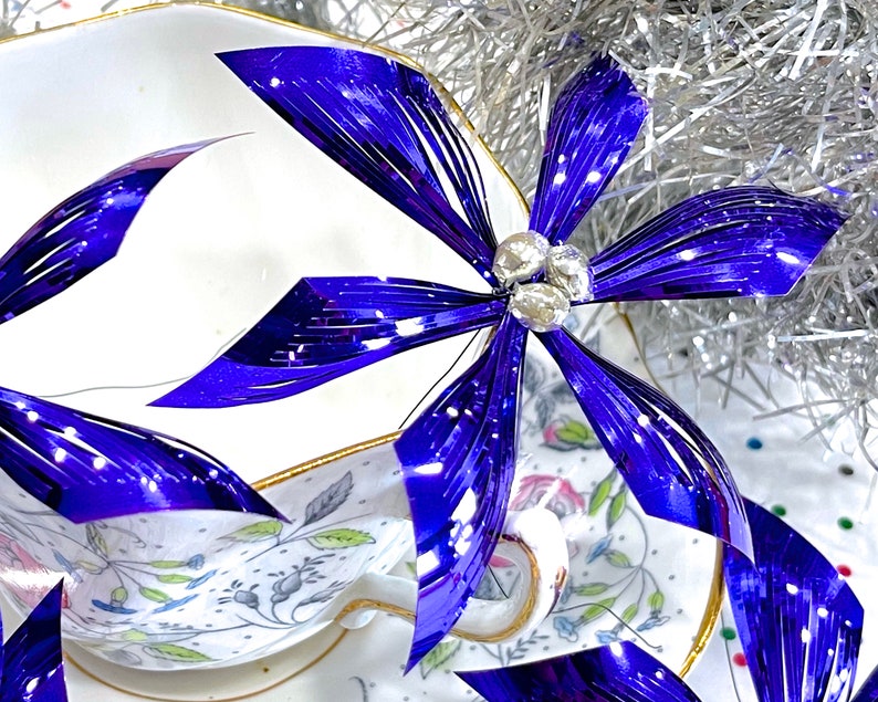 Pu&ograve; includere: Primo piano di un fiocco metallico blu e argento con una tazza da t&egrave; e un piattino bianchi sullo sfondo. Il fiocco &egrave; fatto di strisce sottili e lucide di materiale e ha un aspetto delicato ed elegante.