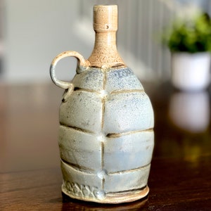 May include: A ceramic bottle with a light blue glaze and a textured surface. The bottle has a handle and a narrow neck.