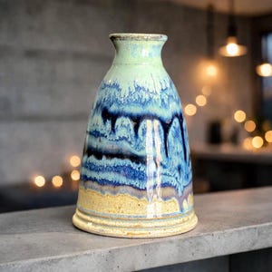 May include: A ceramic vase with a tapered neck and a wide base. The glaze features a gradient transition from a pale lime green at the top, flowing into deep blue and indigo drips in the center, and ending in a textured sandy beige at the bottom.