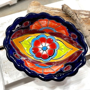 Könnte beinhalten: Eine blaue Keramikschüssel mit gewelltem Rand zeigt ein farbenfrohes Blumenmuster. Die Schüssel hat eine große gelbe, grüne und blaue Blume in der Mitte mit kleineren roten und orangefarbenen Blumen am Rand.