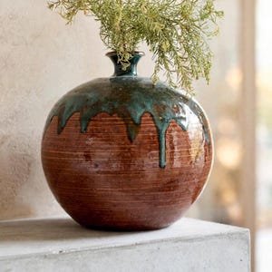 May include: A round ceramic vase with a narrow neck, featuring a textured brown body with horizontal lines and a glossy teal glaze dripping from the rim. The vase contains a sprig of green foliage extending from the top opening.
