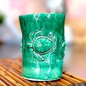 May include: A green ceramic mug with a textured surface and a raised design of sea turtles. The mug is sitting on a bamboo mat.