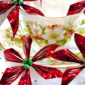 Pu&ograve; includere: Una tazza da t&egrave; bianca e un piattino con un motivo floreale, decorati con fiocchi metallici rossi e verdi. I fiocchi sono realizzati in un materiale lucido e stropicciato.