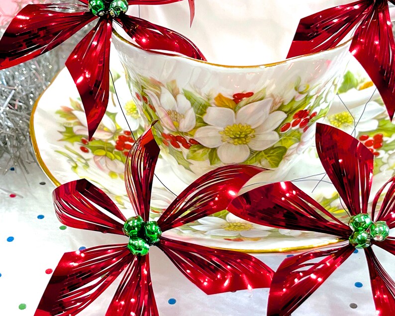Pu&ograve; includere: Una tazza da t&egrave; bianca e un piattino con un motivo floreale, decorati con fiocchi metallici rossi e verdi. I fiocchi sono realizzati in un materiale lucido e stropicciato.