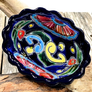 Pode incluir: Um pequeno prato de cerâmica oval com uma borda ondulada. O prato é pintado com um esmalte azul vibrante com um desenho floral em tons de vermelho, amarelo, verde e azul.