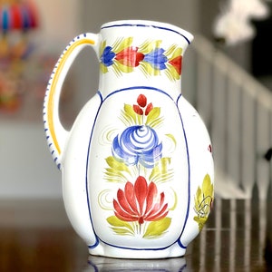 May include: A white ceramic pitcher with a yellow handle and blue, red, and yellow floral designs.