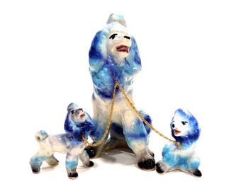 May include: Three porcelain poodle figurines, one large and two smaller, are painted in shades of blue and white. The larger poodle is standing and has a gold chain attached to its collar. The two smaller poodles are sitting and are connected to the larger poodle by the gold chain.