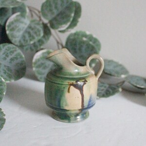 May include: A small, green, blue, and brown ceramic pitcher with a handle. The pitcher is sitting on a white surface.