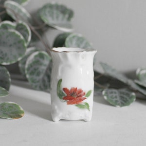 Könnte beinhalten: Eine kleine weiße Porzellanvase mit einer roten Blume, die auf die Vorderseite gemalt ist. Die Vase hat einen gewellten Rand und einen goldenen Rand. Sie steht auf einer weißen Oberfläche.