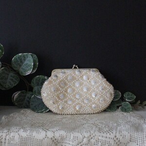 May include: A white beaded clutch purse with a silver clasp. The purse is sitting on a white crocheted doily.