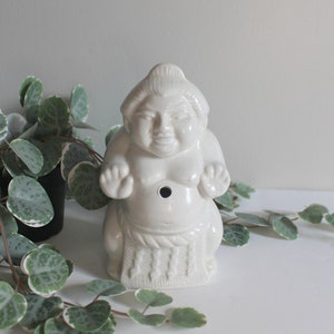 May include: A white ceramic sumo wrestler figurine with a black hole in the chest. The figurine has a detailed design of a traditional sumo wrestler with a topknot hairstyle and a mawashi. The figurine is on a white surface.