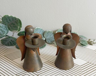 Puede incluir: Dos candelabros de ángel de latón vintage. Cada ángel tiene un cuerpo cónico, pequeñas alas y un portavelas circular. Los ángeles se exhiben sobre una superficie blanca con un patrón geométrico, con vegetación y una cesta de mimbre en el fondo.