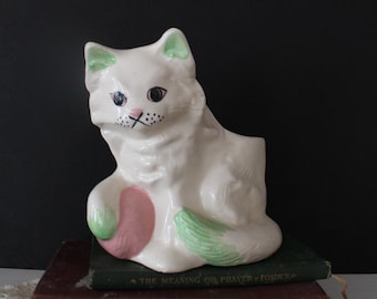 May include: White ceramic cat planter with green ears and a pink flower. The planter is sitting on top of two books, one green and one brown.