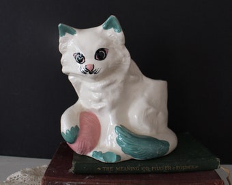May include: White ceramic cat planter with green and pink painted details. The cat is sitting with its tail curled around its body. The planter is sitting on top of two books. The top book has the text "The Meaning of Prayer - Fosdick" printed on the spine.