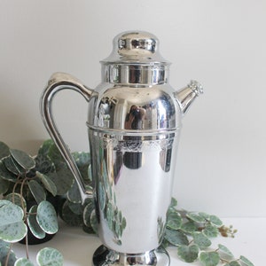 Puede incluir: Una coctelera de plata con una banda decorativa alrededor del centro y un pico en el lateral. La coctelera tiene un asa y una base redonda.