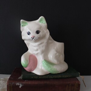 Pode incluir: Vaso de cerâmica branco em forma de gato com orelhas verdes e uma flor rosa. O vaso está em cima de dois livros, um verde e um marrom.