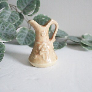 May include: A small, cream-colored ceramic pitcher with a floral design. The pitcher has a rounded body and a short spout. It is decorated with raised flowers and leaves.