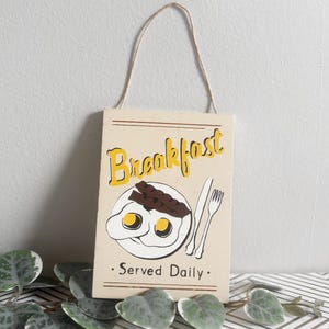 May include: A rectangular wooden sign with the word "Breakfast" in yellow script. The sign features a plate with eggs and bacon, a fork, and a knife. The text "Served Daily" is below the illustration. A beige string is attached for hanging.