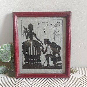 Puede incluir: Arte de silueta enmarcado que representa a un hombre arrodillado ante una mujer. Las figuras en blanco y negro se encuentran sobre un fondo texturizado, enmarcado en un marco de madera rojo desgastado. La obra representa una escena rom&aacute;ntica.
