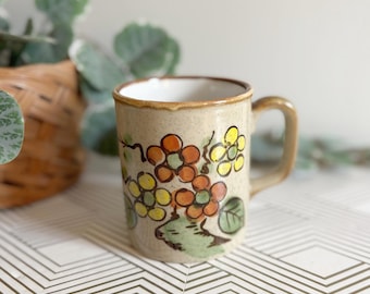 Puede incluir: Una taza de cerámica beige moteada con un diseño floral pintado a mano en tonos naranja, amarillo y verde. La taza tiene un cuerpo cilíndrico, un borde marrón y un asa curva en el lado derecho, con un acabado interior de color blanco.
