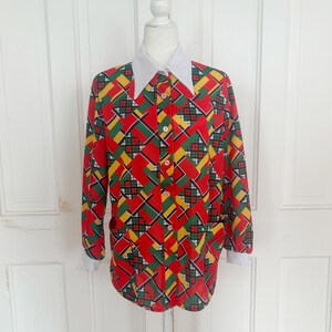 May include: A vintage button-down shirt with a bold geometric pattern in red, yellow, green, and black. The shirt features a white collar and cuffs, and is displayed on a mannequin. The shirt has long sleeves and a classic fit.