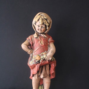May include: A vintage ceramic figurine of a girl wearing a red dress and a white bonnet, holding a basket of fruit. The figurine is standing on a small base.