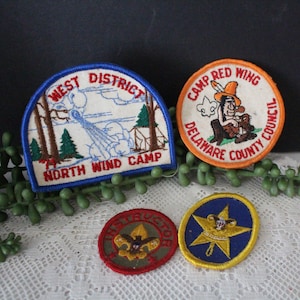 May include: Three vintage Boy Scout patches. One is blue and white with the text "West District North Wind Camp". Another is orange and white with the text "Camp Red Wing Delaware County Council". The last is red and gold with the text "First Class".