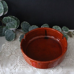 May include: A terracotta ashtray with a brown glaze. The ashtray has a round shape and a slightly raised rim.