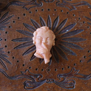 May include: A peach-colored, vintage-style cameo of a person with a finger to their lips, set against a carved wooden background. The cameo is a bust, suggesting silence or secrecy. The wooden surface features floral and ribbon designs.