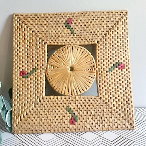 Puede incluir: Un marco cuadrado de mimbre tejido con una pieza central redonda de mimbre tejido. El marco tiene un diseño decorativo de flores rojas y verdes.