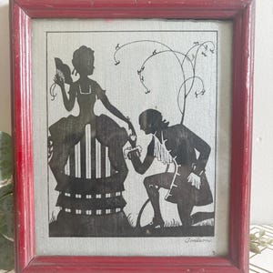 Puede incluir: Arte de silueta enmarcado que representa a un hombre arrodillado ante una mujer, ambos con atuendos de &eacute;poca. Las siluetas negras se colocan sobre un fondo plateado, enmarcadas en madera roja envejecida. La obra de arte representa una escena rom&aacute;ntica.