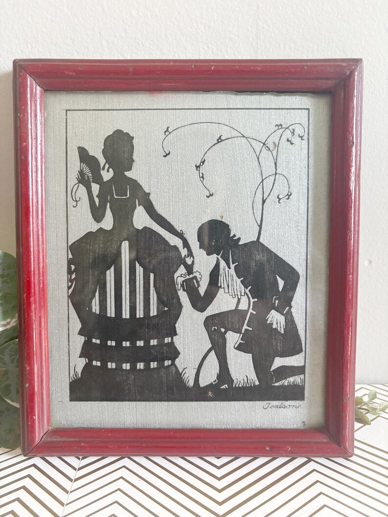 Puede incluir: Arte de silueta enmarcado que representa a un hombre arrodillado ante una mujer, ambos con atuendos de &eacute;poca. Las siluetas negras se colocan sobre un fondo plateado, enmarcadas en madera roja envejecida. La obra de arte representa una escena rom&aacute;ntica.
