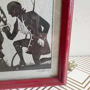 Puede incluir: Arte de silueta enmarcado que representa una figura negra con vestimenta de &eacute;poca, arrodillada sobre hierba. La obra de arte est&aacute; sobre un fondo plateado texturizado y enmarcada en un marco de madera rojo. La firma del artista, "Joalsons", es visible.