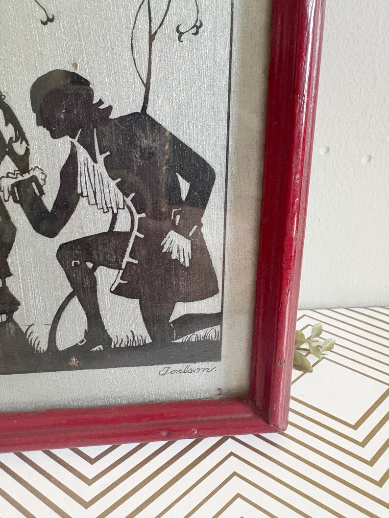Puede incluir: Arte de silueta enmarcado que representa una figura negra con vestimenta de &eacute;poca, arrodillada sobre hierba. La obra de arte est&aacute; sobre un fondo plateado texturizado y enmarcada en un marco de madera rojo. La firma del artista, "Joalsons", es visible.