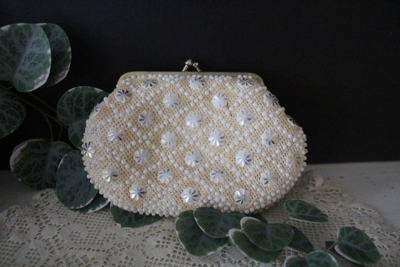 May include: A white beaded clutch purse with a silver floral pattern. The purse has a clasp closure.