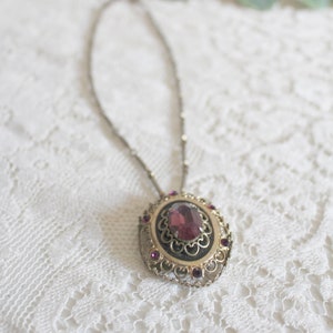 May include: A silver chain necklace with a pendant featuring a large, oval-shaped, purple gemstone surrounded by a gold filigree design and smaller purple gemstones.